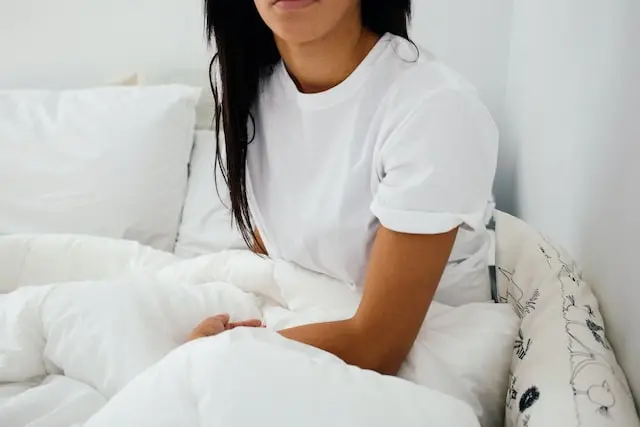 A woman with long hair sits up in bed while still under the covers